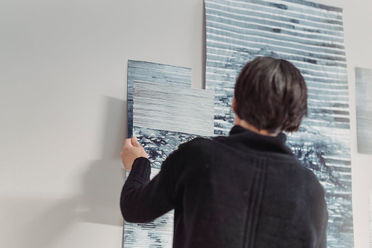 A woman in a dark sweater arranging abstract paintings on a wall, showcasing creative expression.