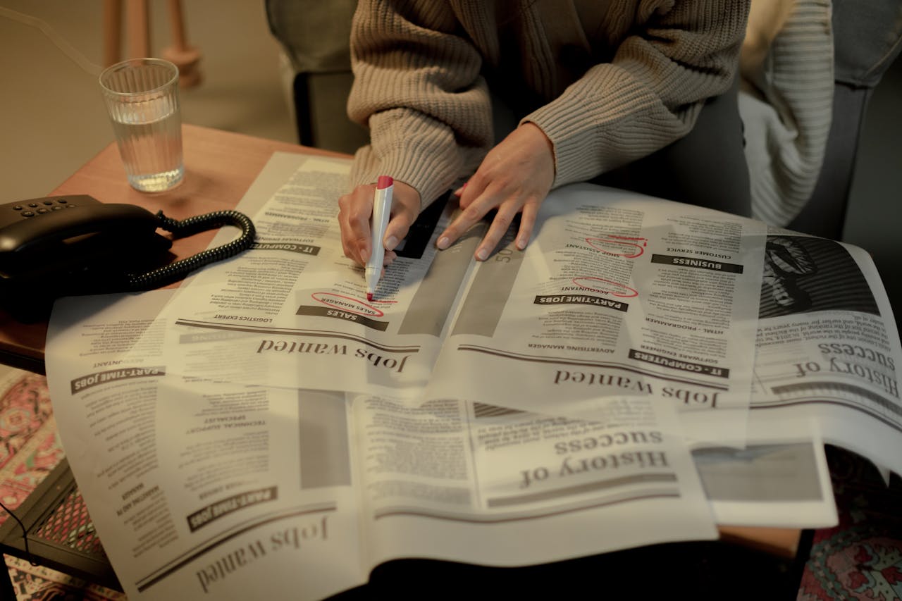 team-02 An adult woman marks job listings in a newspaper while sitting indoors, highlighting job search activity.